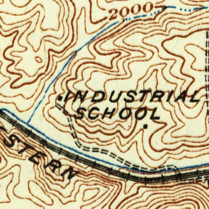 Detail of a 1930s topographic map in brown contour lines: the words Industrial School printed across a hill in the center, a double dashed farm lane winding down from the left of the words to the bottom, a blue stream and a railroad drawn with cross ties running along the bottom, the letters STERN of the railroad’s name at lower left, and a contour labeled 2000 at the top.
