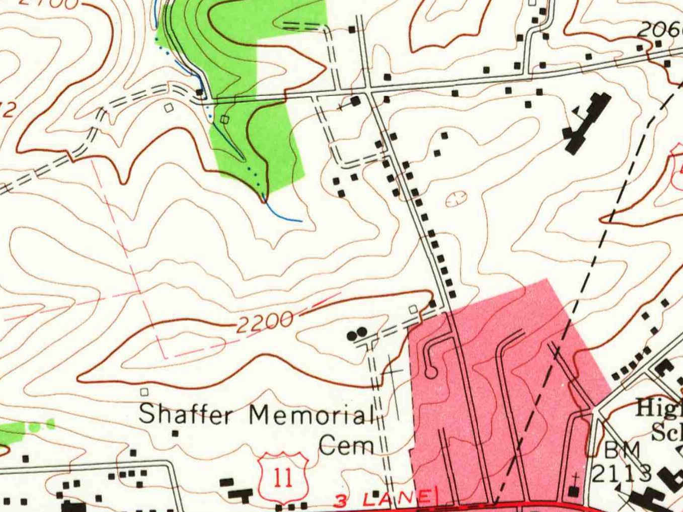 Detail of a 1965 topographic map: open ground with brown contour lines on a hillside, a small stream in a strip of green woods at top center, a street lined with black house squares running down the right side to a road marked 11, the label Shaffer Memorial Cem near the bottom, and the pink built-up area of town at lower right.